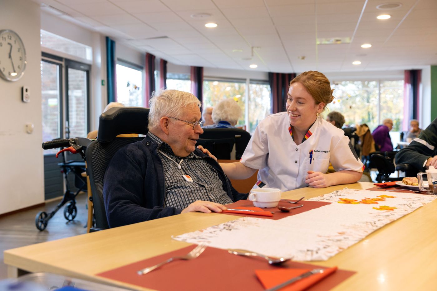 Zorgmedewerker en bewoner in verpleeghuis Het Nieuwe Feithenhof op de Veluwe van christelijke zorgorganisatie Lelie zorggroep verpleeghuiszorg uniform vacature Jonge vrouw in zorgjasje en oude man in rolstoel zitten samen aan tafel in de ontmoetingsruimte van een verpleeghuis