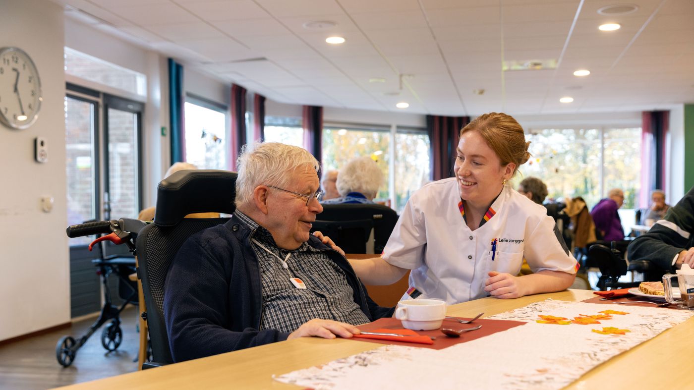Zorgmedewerker en bewoner in verpleeghuis Het Nieuwe Feithenhof op de Veluwe van christelijke zorgorganisatie Lelie zorggroep verpleeghuiszorg uniform vacature Jonge vrouw in zorgjasje en oude man in rolstoel zitten samen aan tafel in de ontmoetingsruimte van een verpleeghuis