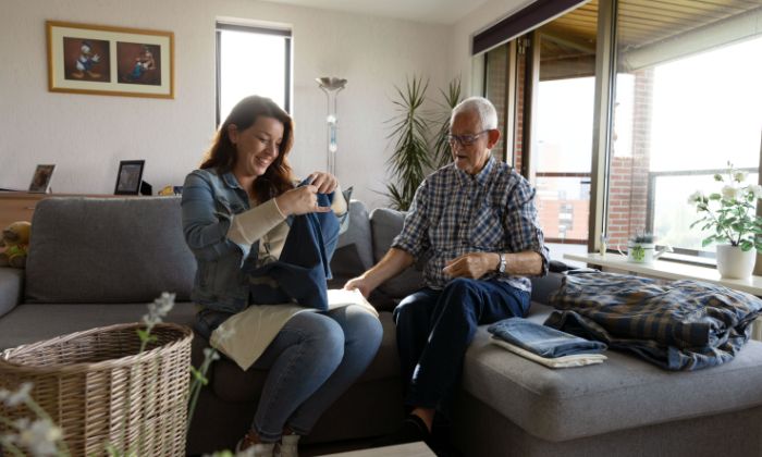 Client en hulp bij huishouden van Lelie zorggroep lachen samen was vouwen Vrouwelijke medewerker hulp bij huishouden vouwt de was op samen met een mannelijke client in een huiskamer op de bank