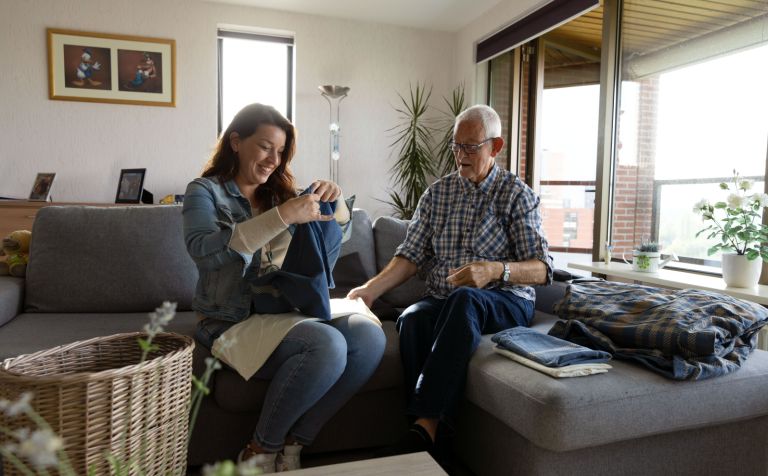 Client en hulp bij huishouden van Lelie zorggroep lachen samen was vouwen Vrouwelijke medewerker hulp bij huishouden vouwt de was op samen met een mannelijke client in een huiskamer op de bank
